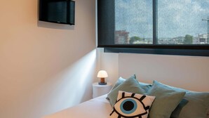 Un dormitorio con una cama, un televisor en la pared, una mesita de noche con lámpara y una ventana con vistas a la ciudad.