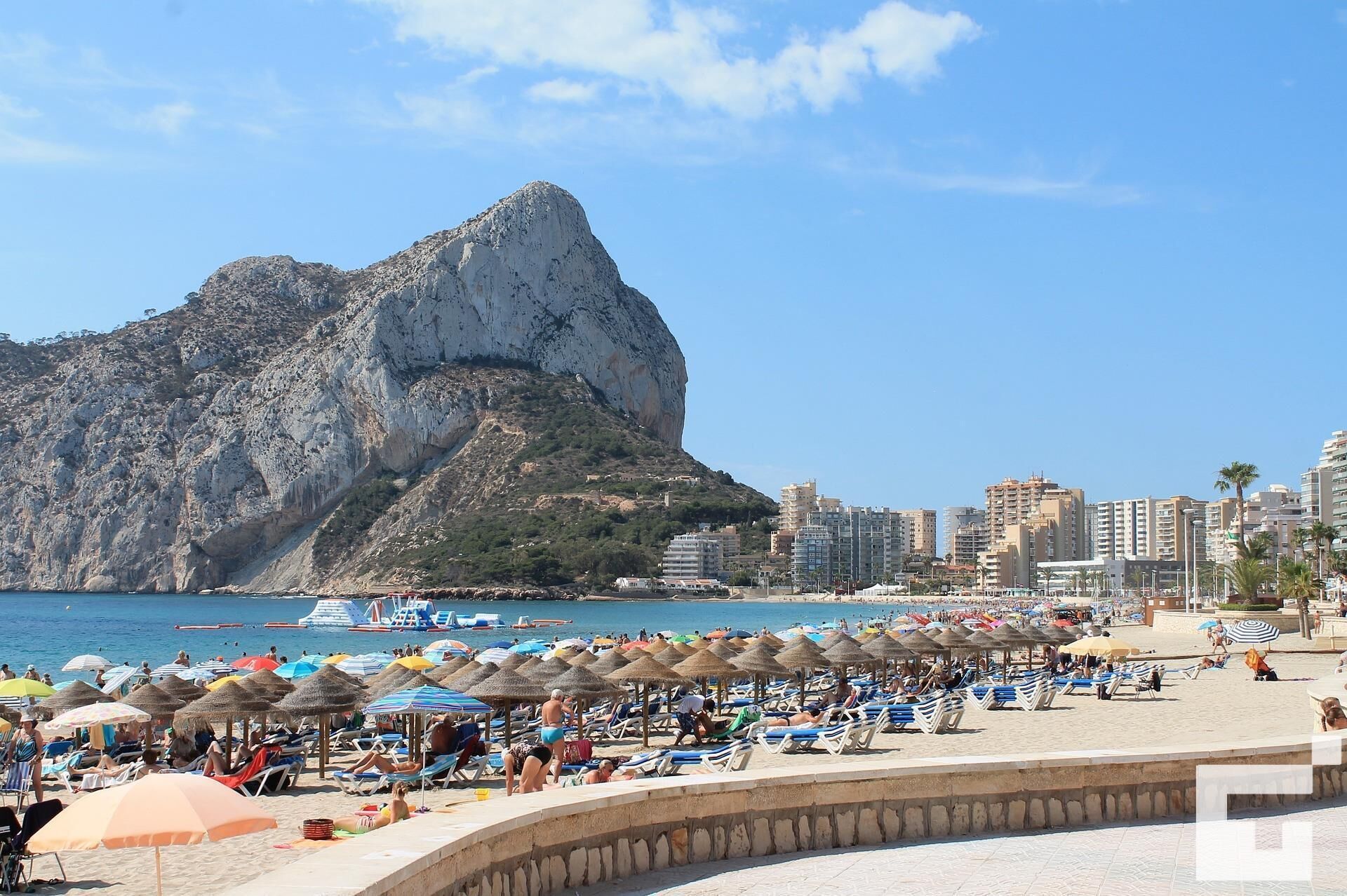 Ein Strand mit Liegestühlen und Sonnenschirmen, ein felsiger Berg im Hintergrund und eine Skyline mit Gebäuden.
