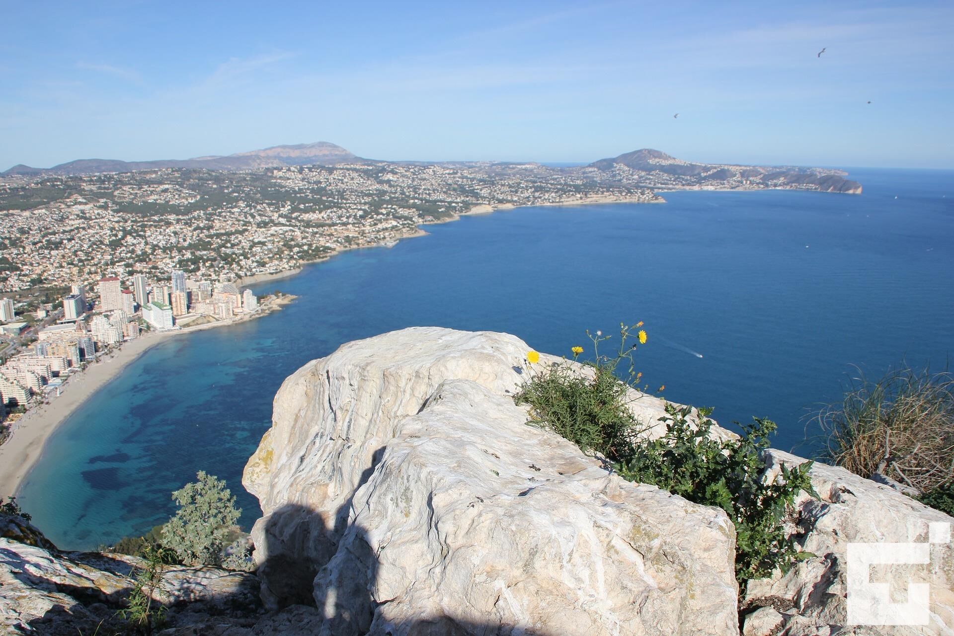 A coastal cityscape with a prominent rocky outcrop in the foreground, a beach, and a view of the city and sea in the background.