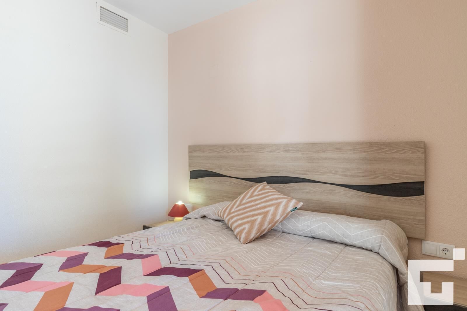 Een slaapkamer met een bed, nachtkastje, lamp en een stopcontact.