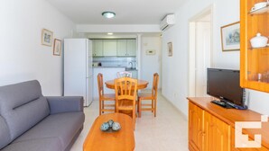 A compact living space with a sofa, a dining table with chairs, a TV on a wooden stand, and a kitchenette in the background. - Zafiro 211B - Grupo Turis - Calpe, Costa Blanca (Calpe)