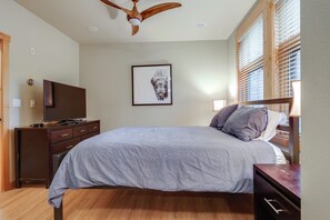 Une chambre à coucher comprenant un lit, une commode, un téléviseur, un ventilateur de plafond et une fenêtre avec des stores.