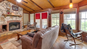 A cozy living room with a stone fireplace, leather sofa, wooden coffee table, and a rocking chair. - Mountaintop Ellijay Cabin w/ Private Hot Tub (Ellijay)