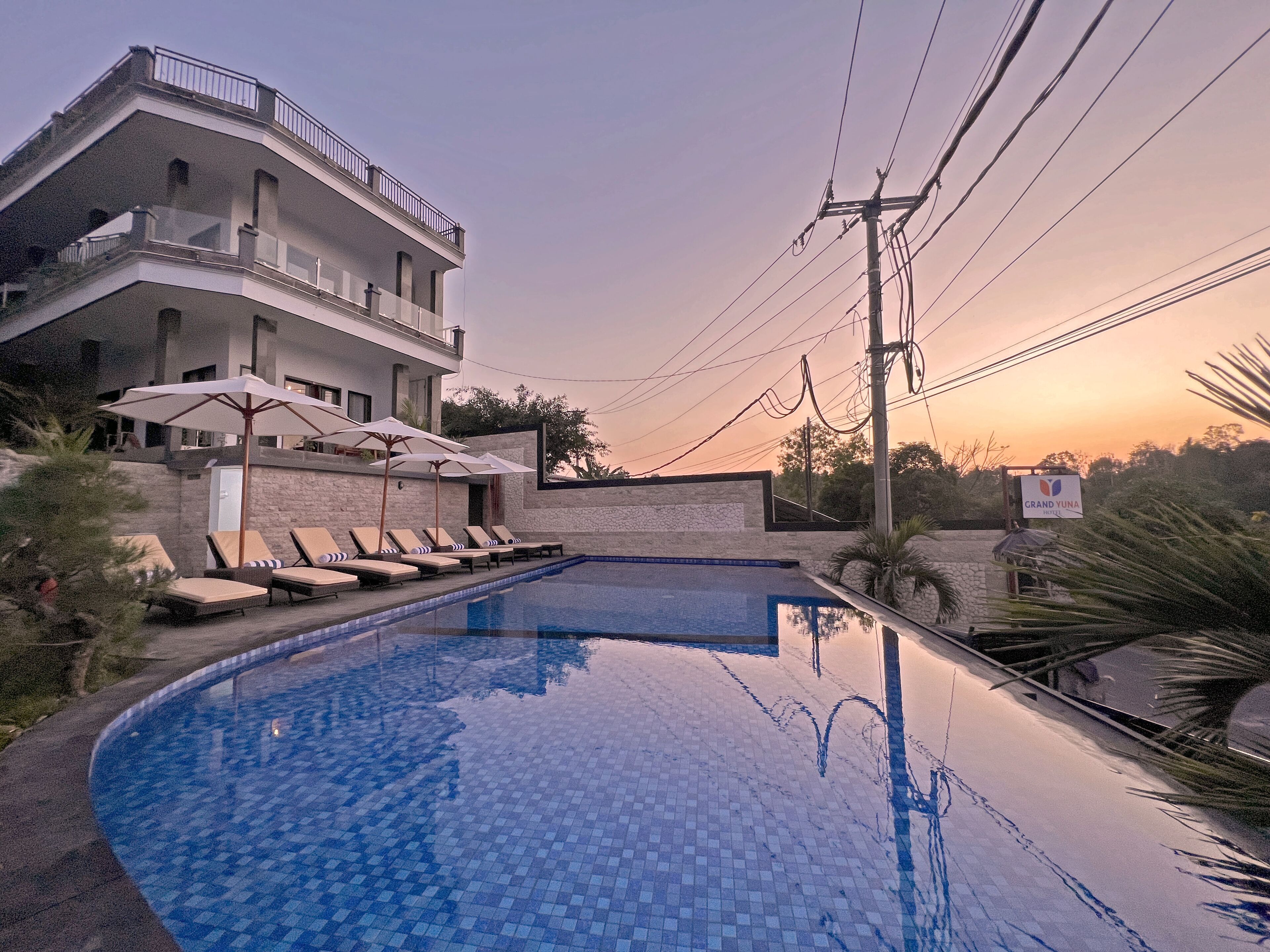 Ein Schwimmbecken mit Liegestühlen und Sonnenschirmen, ein mehrstöckiges Gebäude mit Balkonen und ein Sonnenuntergang im Hintergrund.