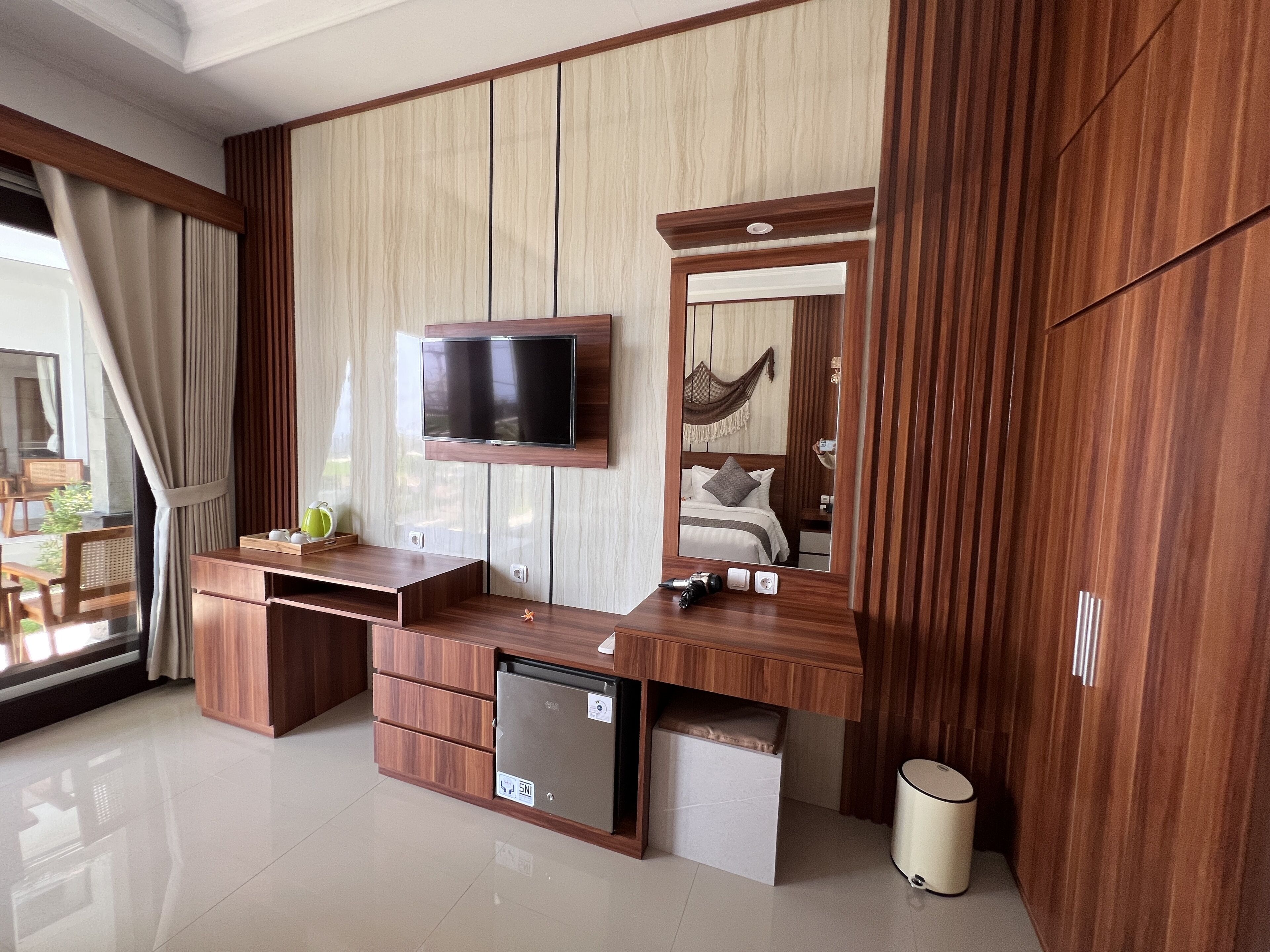 Un dormitorio moderno con un tocador de madera, un televisor de pantalla plana montado encima y un armario empotrado, además de un refrigerador pequeño.