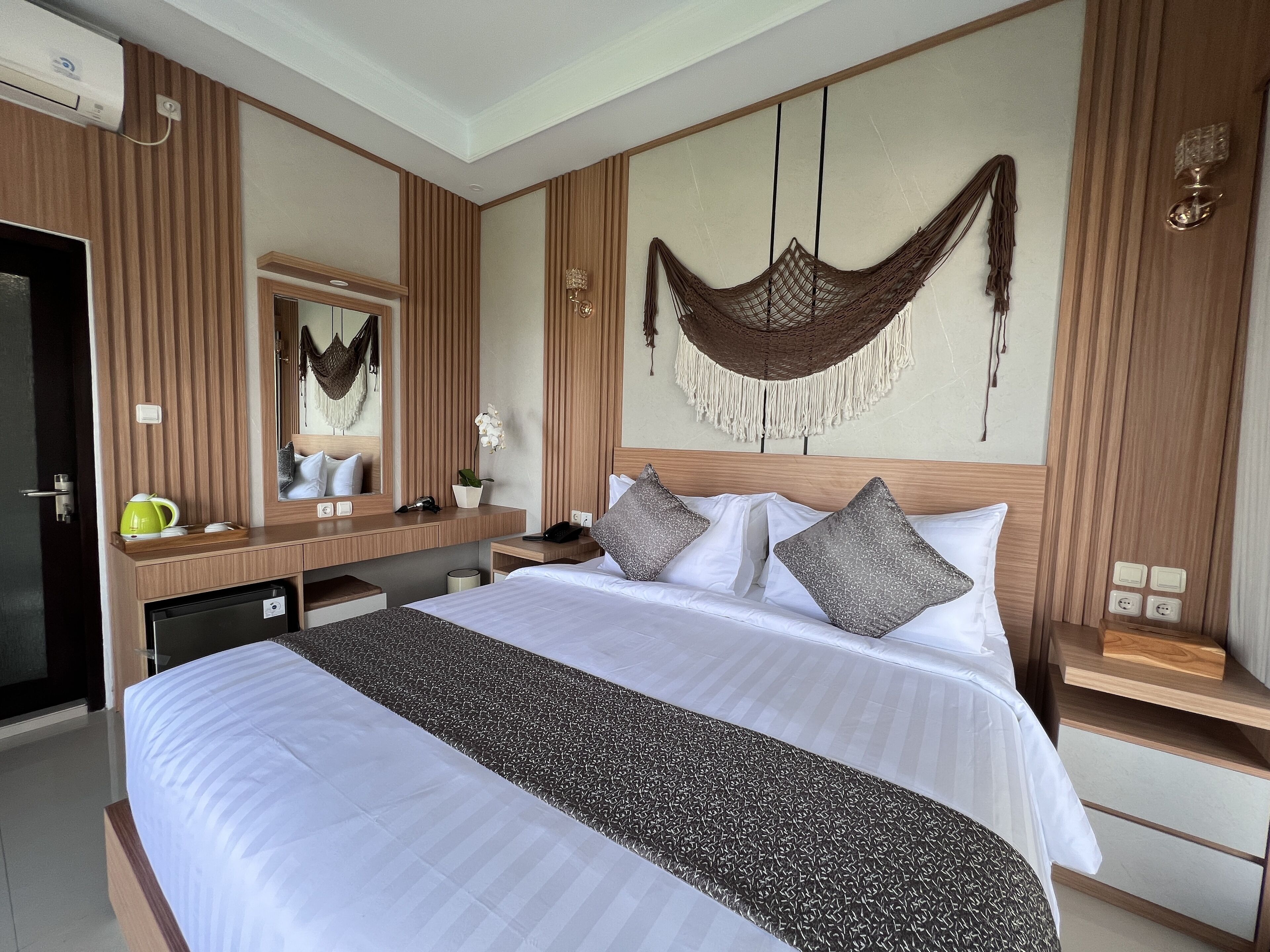 Une chambre d’hôtel avec un grand lit, une tête de lit en bois, une table de chevet avec une lampe, un miroir et un hamac suspendu.