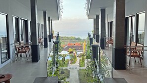 Un balcón moderno de hotel con barandas de vidrio, sillas de madera y vista al mar y un jardín abajo.