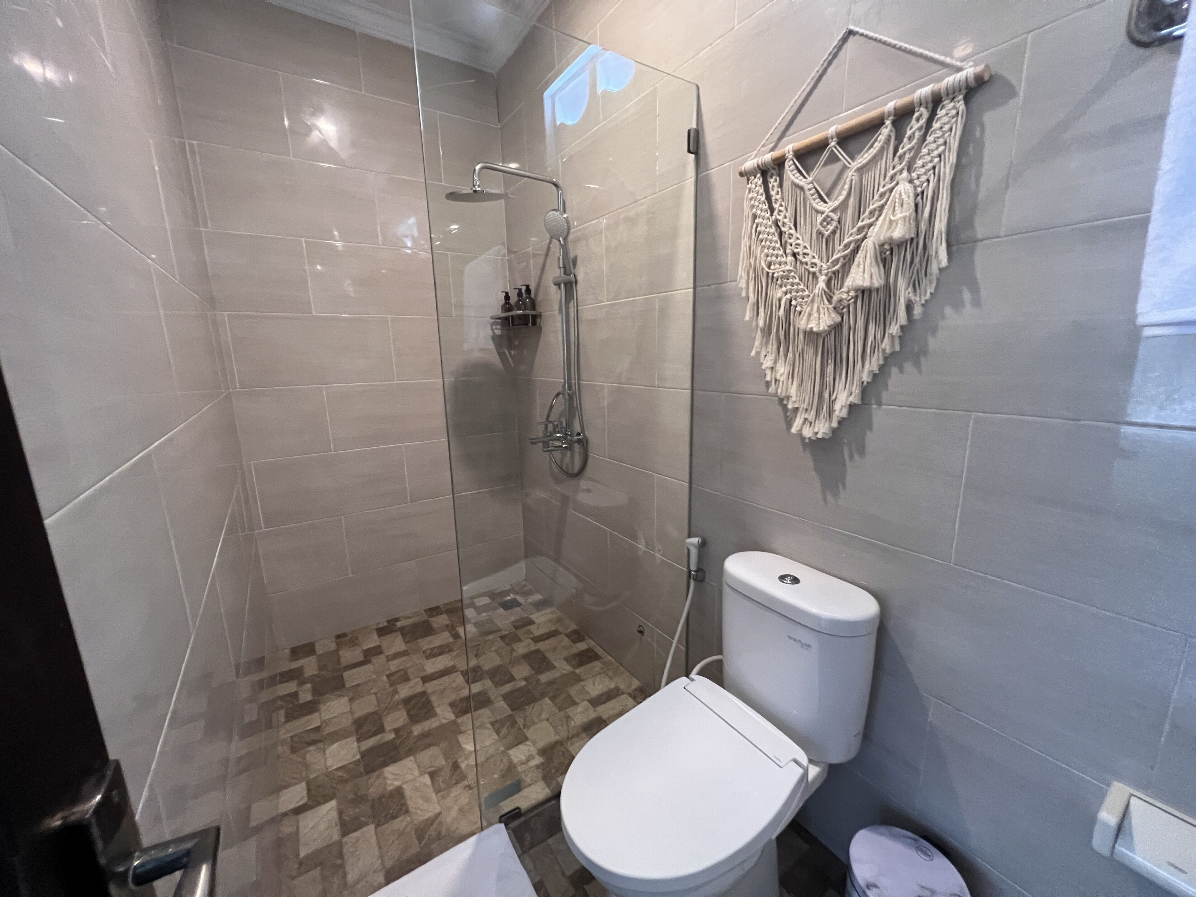 A modern bathroom with a glass-enclosed shower, a toilet, and a macrame wall hanging.