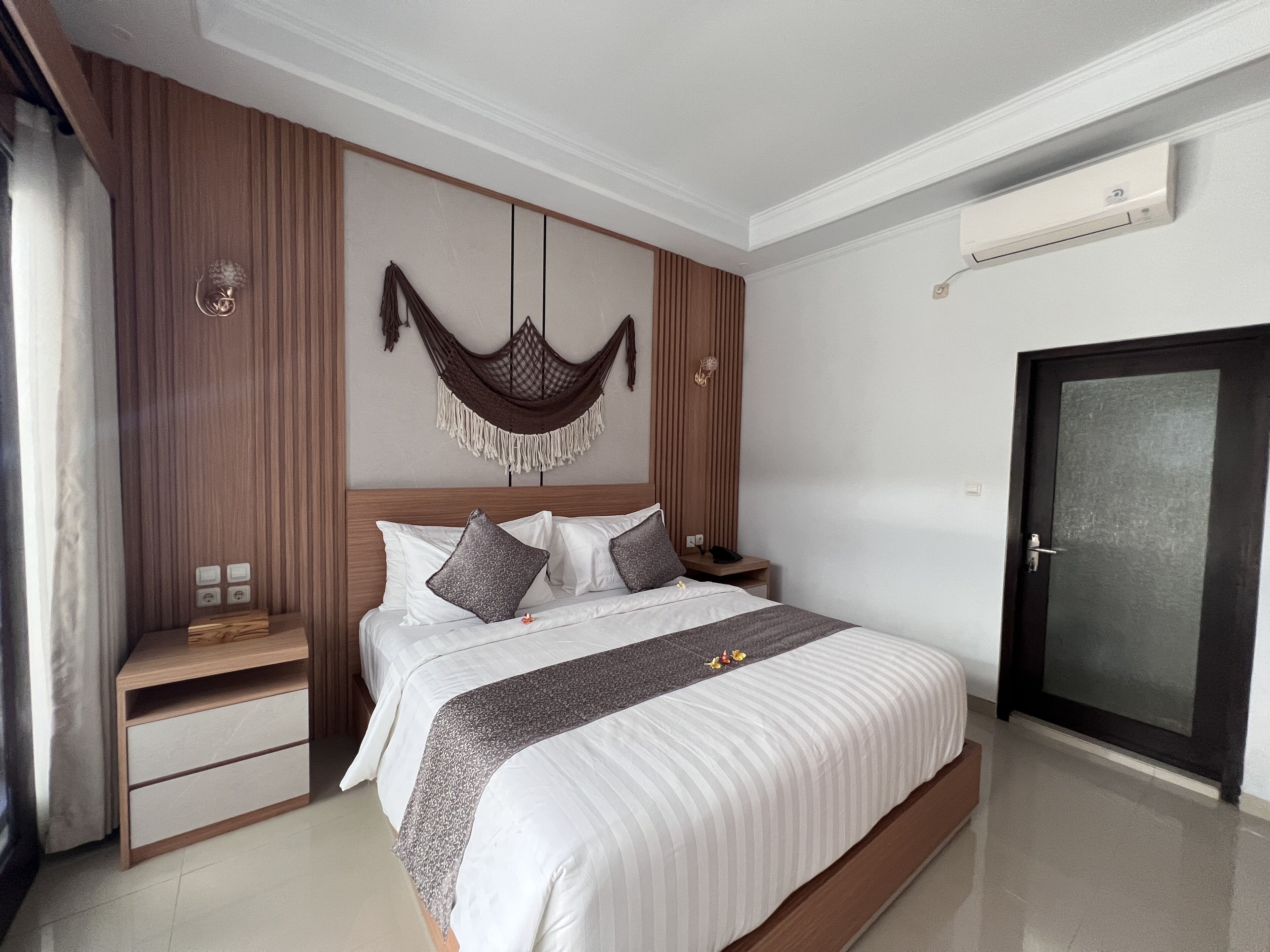 Une chambre moderne avec un grand lit, un lambris en bois sur les murs, un élément décoratif suspendu, une table de chevet avec une lampe et une porte dotée d’un panneau en verre dépoli.