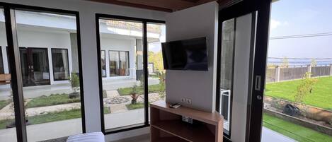 A modern room with a bed, a TV on a wooden stand, and large sliding glass doors leading to a balcony with a view of the sea.