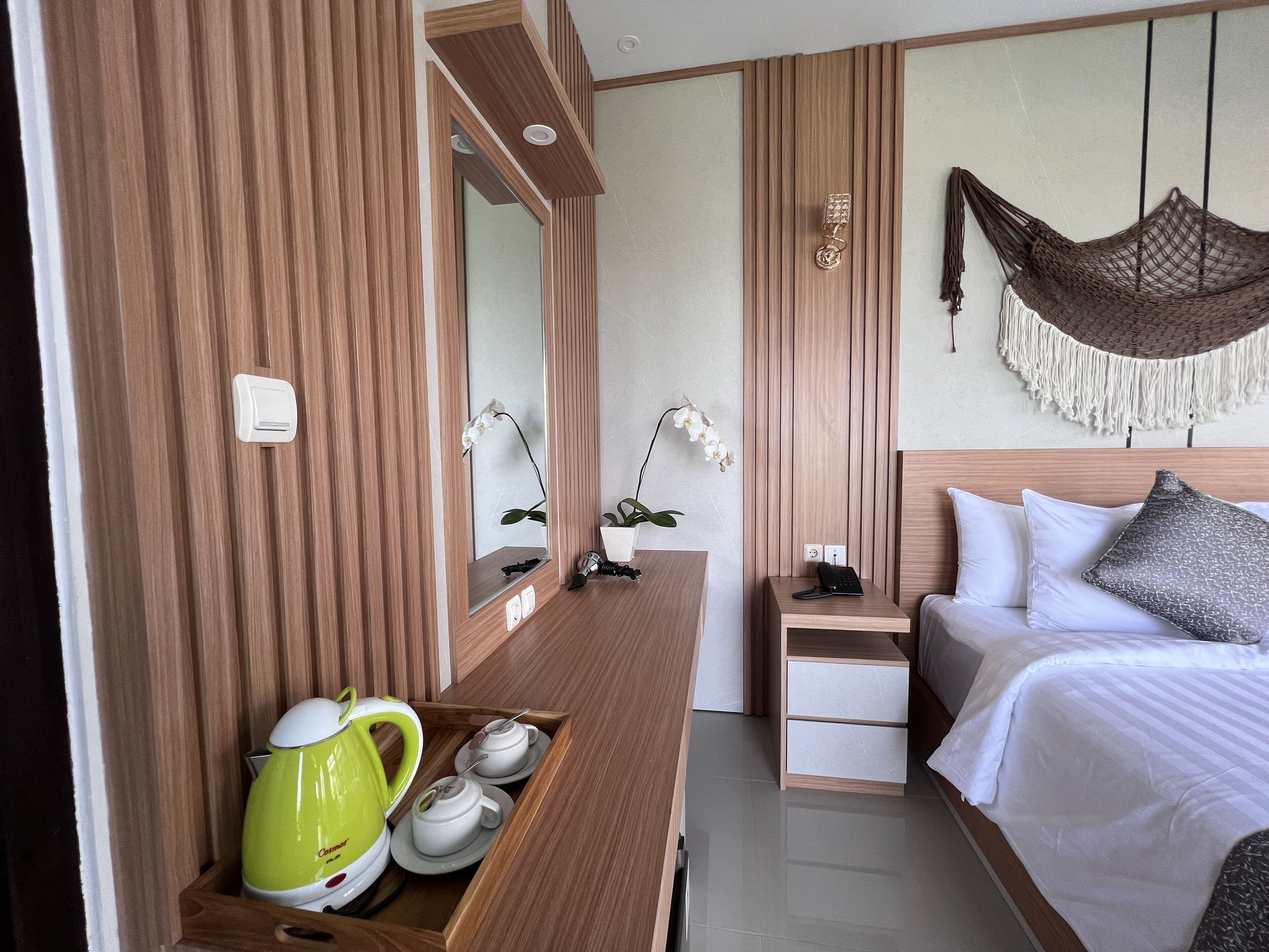 A modern hotel room with a wooden vanity, a bedside table with a phone, a bed with white bedding, and a hanging macrame chair.