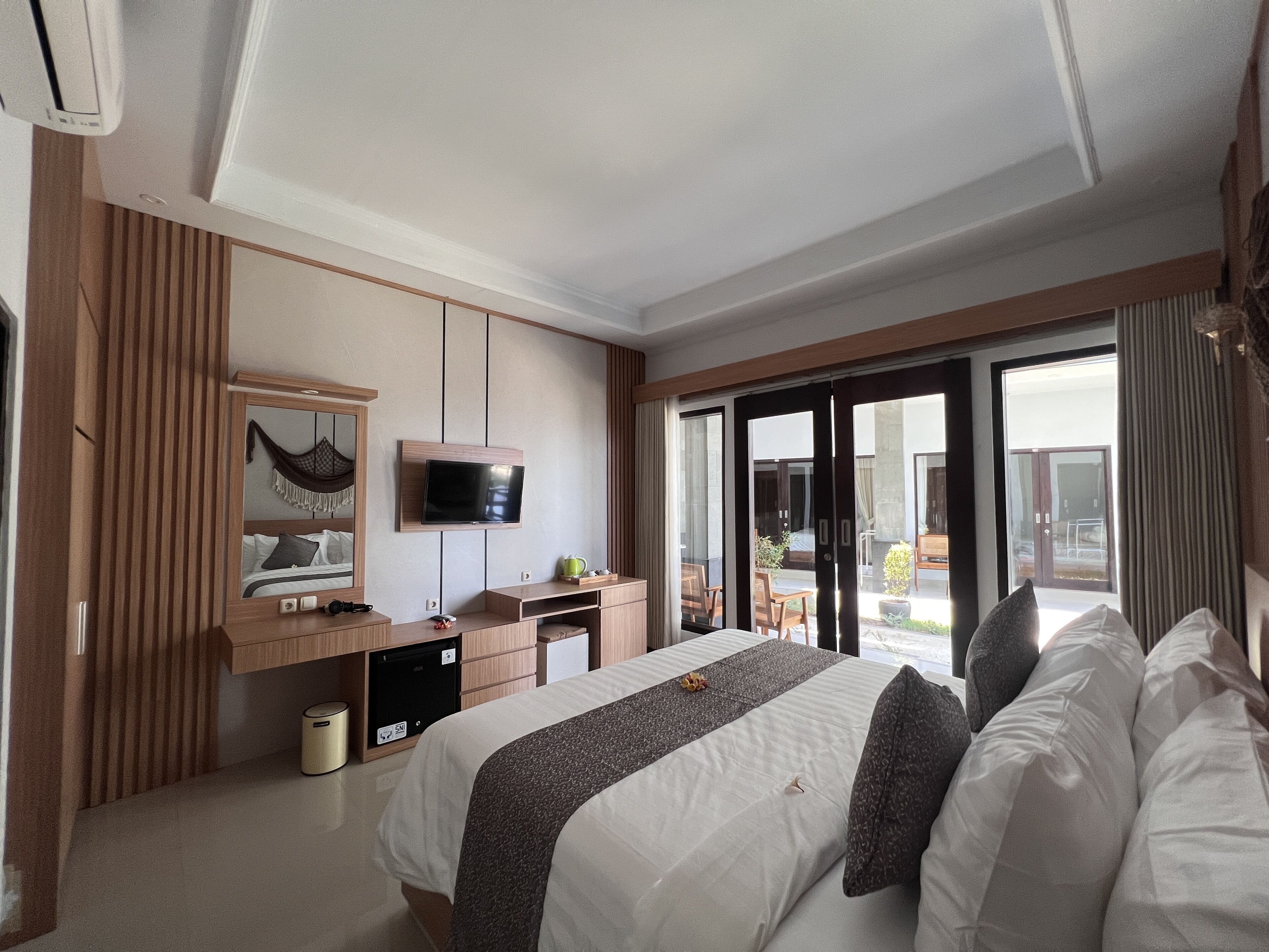 A hotel room with a bed, a desk with a flat-screen TV, a minibar, and a sliding glass door leading to an outdoor area.