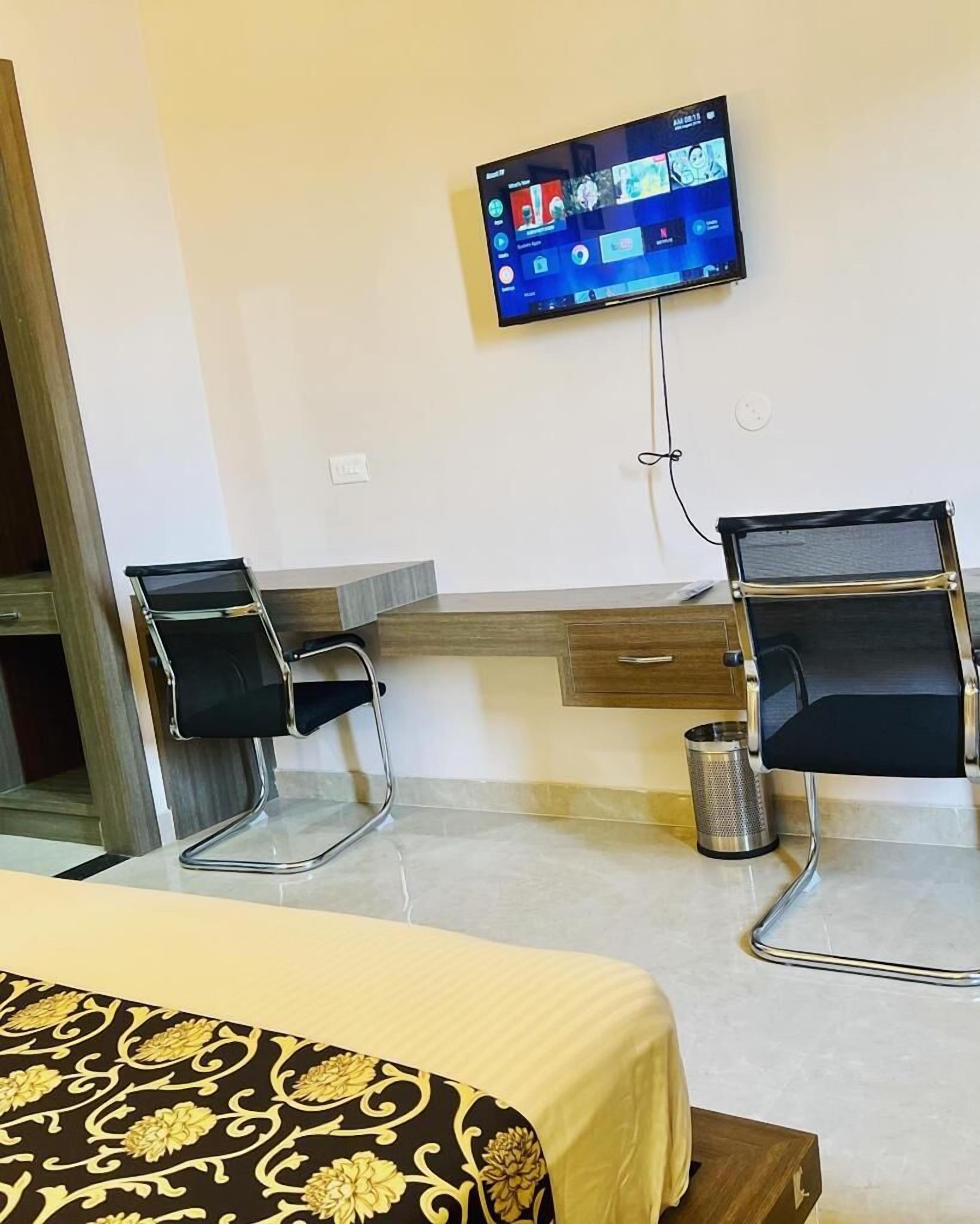 A hotel room with a bed, two chairs, a desk, a television mounted on the wall, and a trash can.