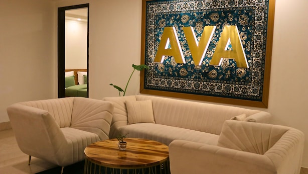 A modern living room with a beige sectional sofa, a wooden coffee table, and a large decorative wall piece with the letters "AVA". - AVA Hotels and Corporate Stays Golf course (Gurugram)