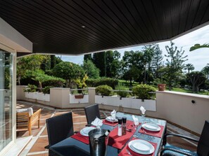 Uma área de refeições ao ar livre com uma mesa posta para quatro pessoas, rodeada de cadeiras, sob um terraço coberto.