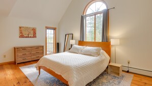 A bedroom with a large bed, a wooden dresser, a window with curtains, a painting on the wall, and a rug. - Charming Putney Home: Porch, Grill & Hiking Trails (Putney)