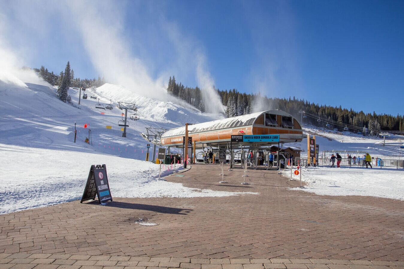 Ein Skigebiet mit überdachter Wartezone, einer Seilbahn im Hintergrund und einem Schild mit der Aufschrift 'Ski & Rider Gondola Load'.