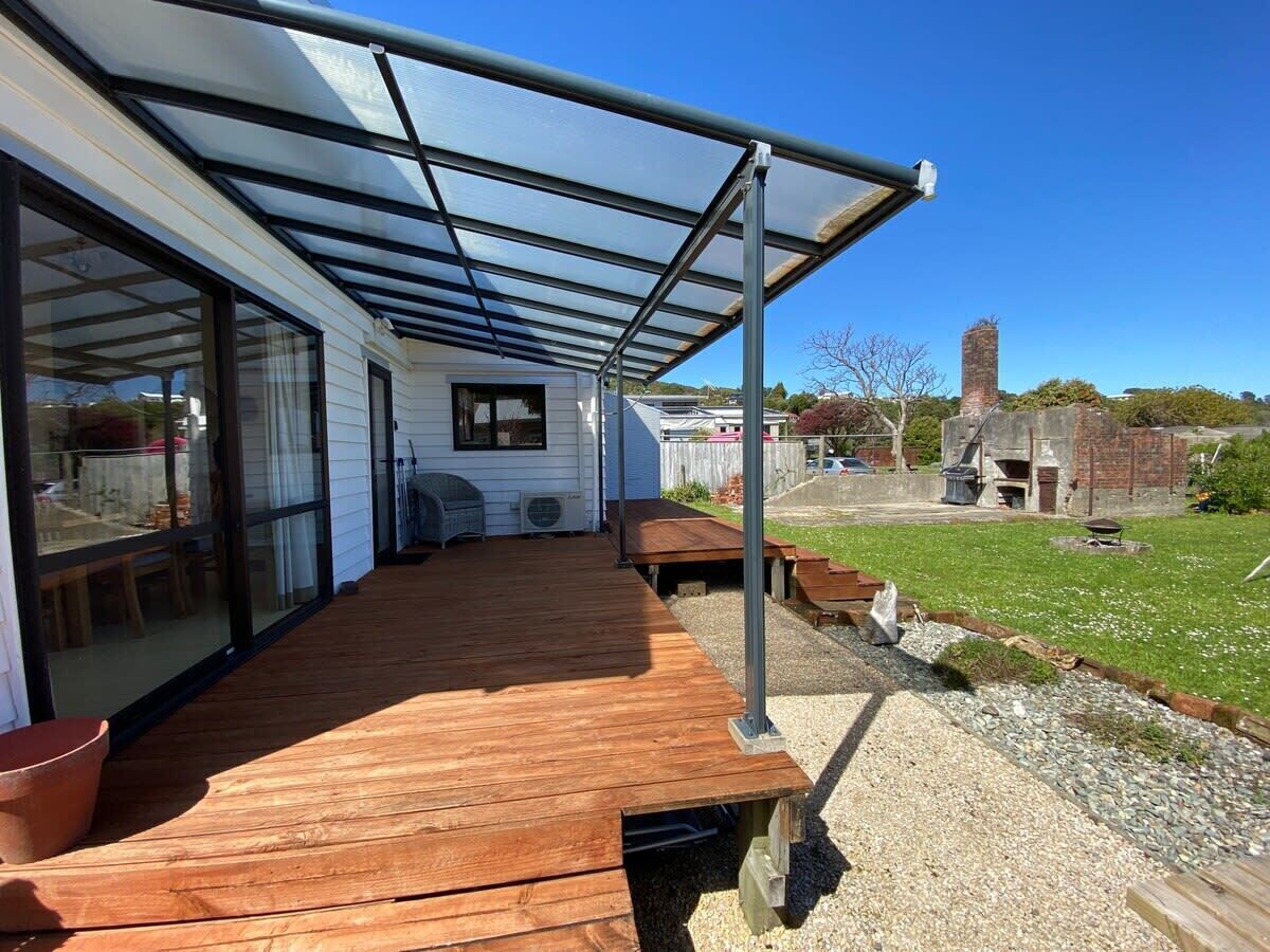 En overdækket terrasse med træterrasse, store skydedøre og udsigt til en baghave med skorsten i mursten og græsplæne.