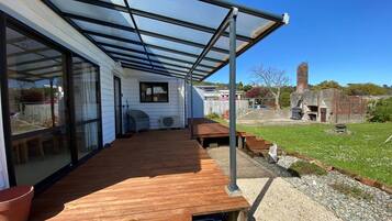 En overdækket terrasse med træterrasse, store skydedøre og udsigt til en baghave med skorsten i mursten og græsplæne.