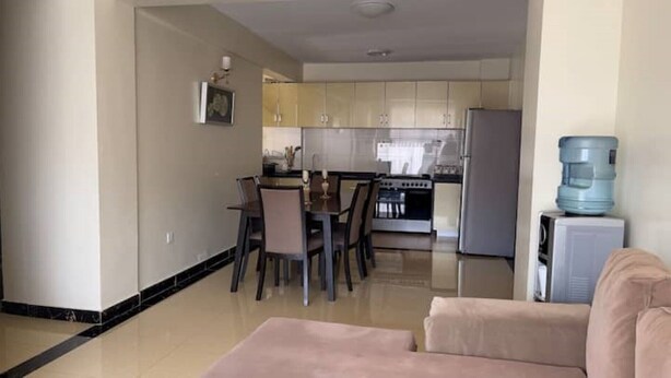 A living room with a beige sofa, a dining area with a table and chairs, a kitchen with cabinets and appliances, and a water dispenser.