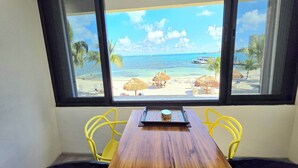 A wooden table with two yellow chairs in front of a window overlooking a beach with thatched umbrellas. - Oceanfront Condo · Hotel Zone · Near Isla Ferry (CancÃºn)
