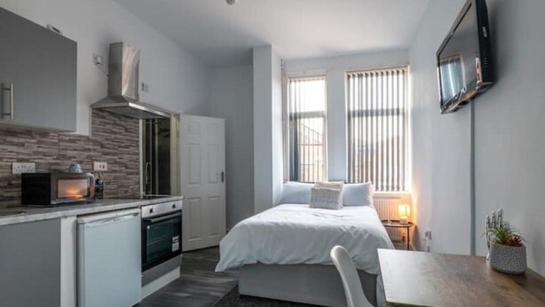 A compact hotel room with a kitchenette, a bed with white bedding, a small table, and a TV mounted on the wall. - Elegant Studio: Studio Suite 5 (Coventry)