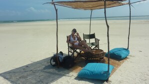 A beach scene with a person sitting under a thatched structure, a wicker chair, and a blue cushion on the sand.