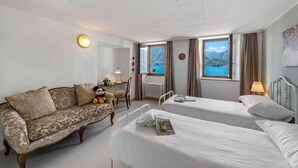 A hotel room with a sofa, two beds, a window with a view of the sea, a lamp, and a radiator. - Casa Matilde Apartments (Gambarogno)
