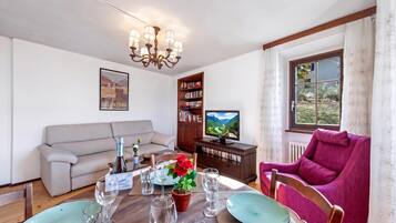 Ein Wohnzimmer mit einer Couch, einem Esstisch für zwei Personen, einem Bücherregal, einem Fernseher und einem Fenster mit Vorhängen.