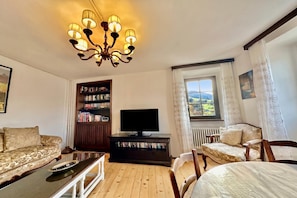 A living room with a chandelier, a sofa, a television on a stand, a bookshelf, and a window with curtains.