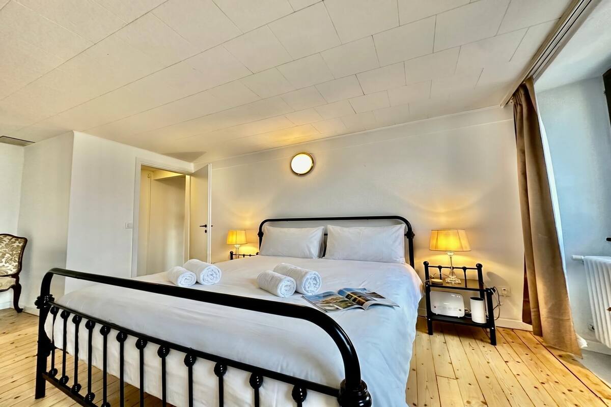 A neatly made bed with rolled towels, a bedside table with a lamp, and a small bedside table with a magazine.