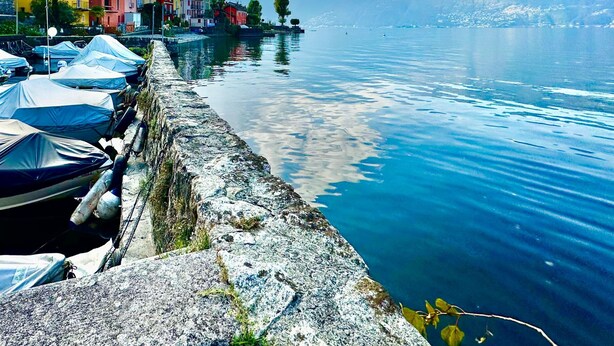A waterfront with colorful buildings, a stone wall, and boats covered with tarps. - Casa Matilde Apartments (Gambarogno)