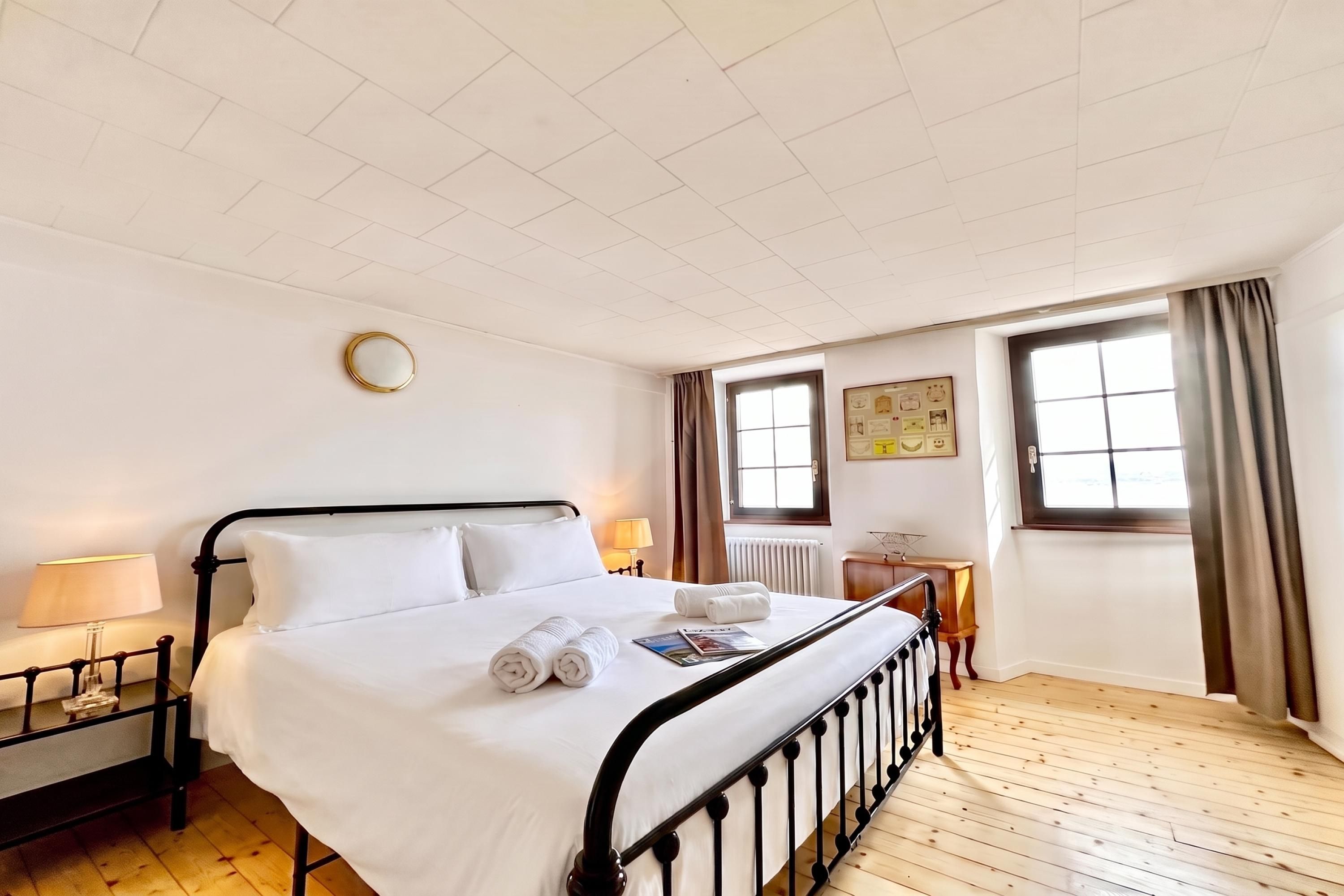 Ein Hotelzimmer mit Bett, Nachttisch, Lampe, einem Fenster mit Vorhängen und einer Wanduhr.