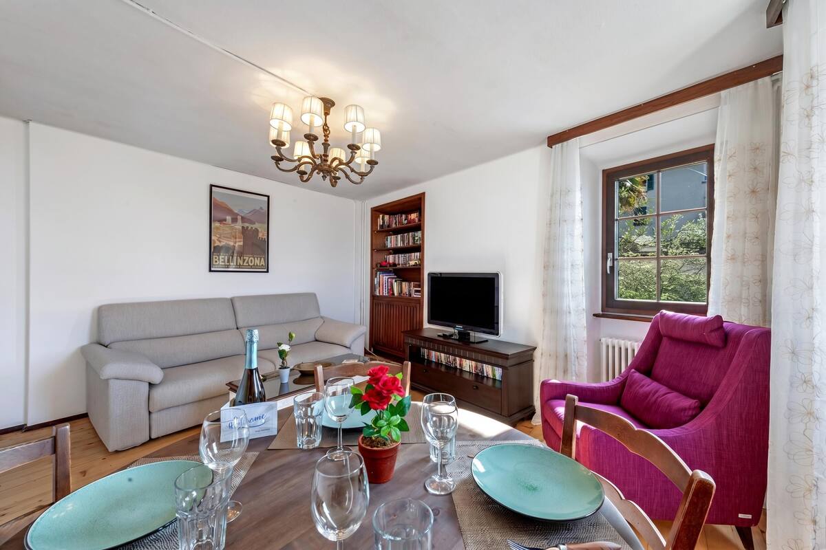 A living room with a sofa, a dining table set with plates and glasses, a TV, a bookshelf, and a window with curtains.