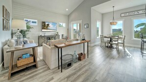 A spacious living area with a wooden table, a sofa, and a dining table with chairs. There are large windows providing natural light and a view of the outdoors.