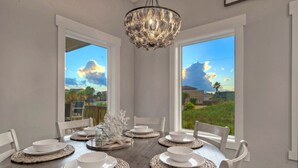 A dining table set with white plates and bowls, surrounded by white chairs, with a chandelier above and large windows offering a view of the outdoors.