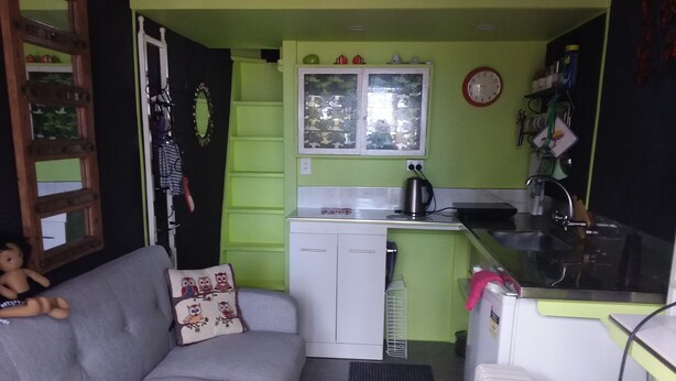 A compact living space with a kitchenette, a grey sofa with a decorative pillow, and a small window with a decorative frame. - TIny House experience, Lake Rotoiti, Rotorua (Rotoiti Forest)