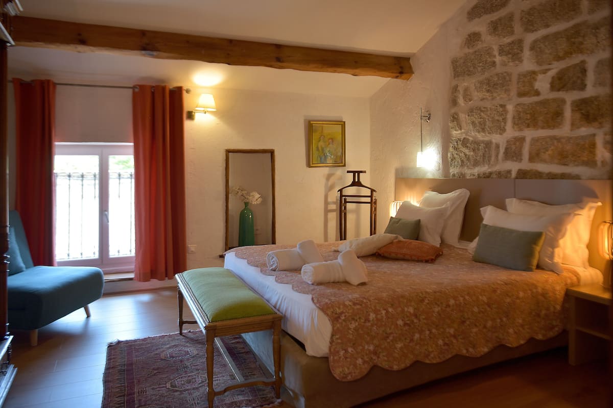 Ein Schlafzimmer mit Bett, einer Bank, einem Sessel, einem Nachttisch, einer Lampe, einem Spiegel und einem Fenster mit Vorhängen.