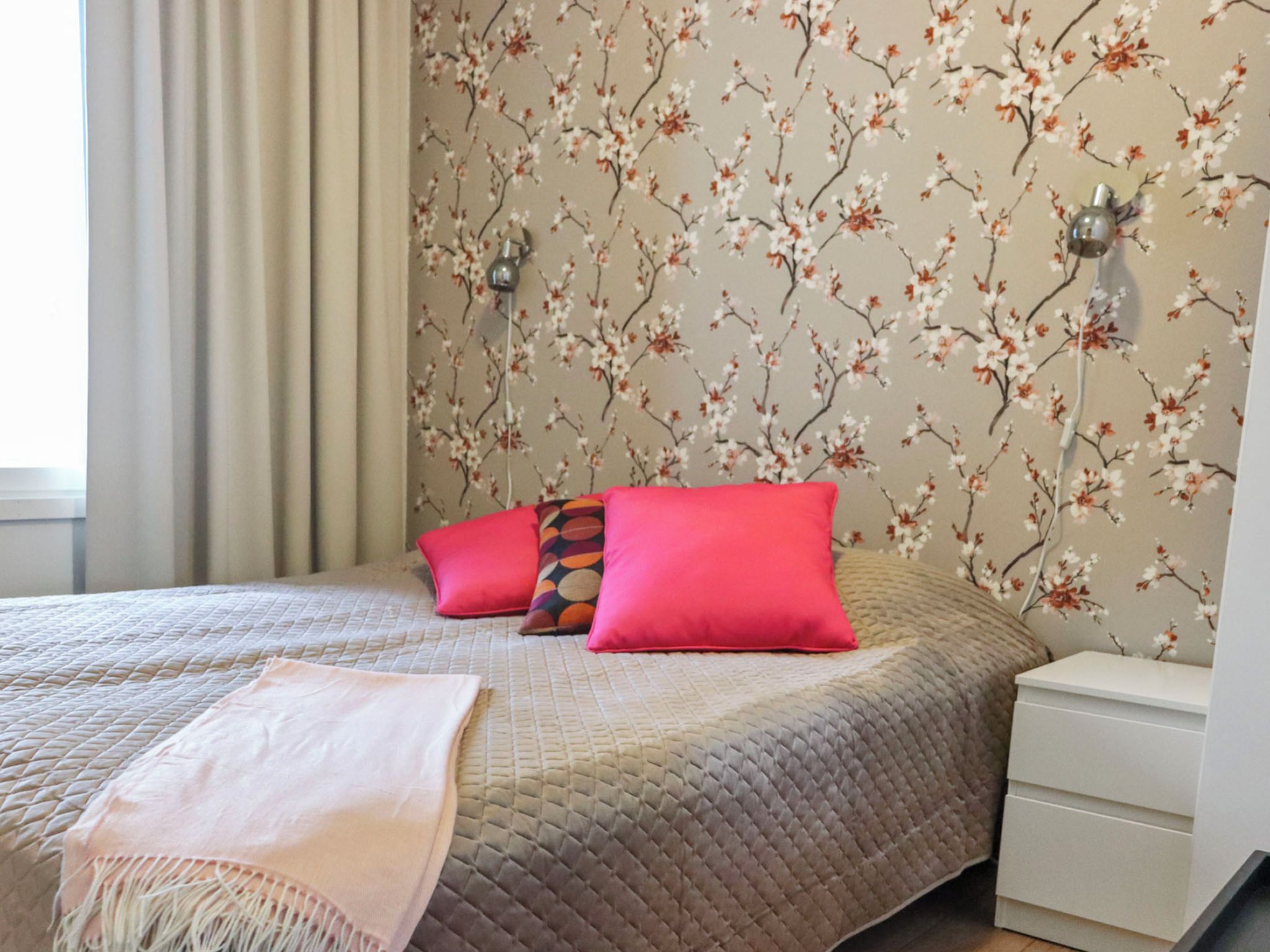 A bedroom with a bed, pillows, a bedside table, and a wall with floral wallpaper.