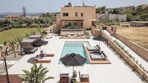 A pool area with lounge chairs and umbrellas, a pool, and a building with a flat roof in the background. - Villa Agali Free* heated pool & 70m to Sea! (Chania)
