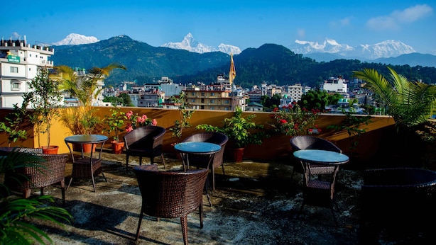 A rooftop terrace with wicker furniture and potted plants, overlooking a cityscape and snow-capped mountains. - Hotel Hidden Kingdom (Pokhara)