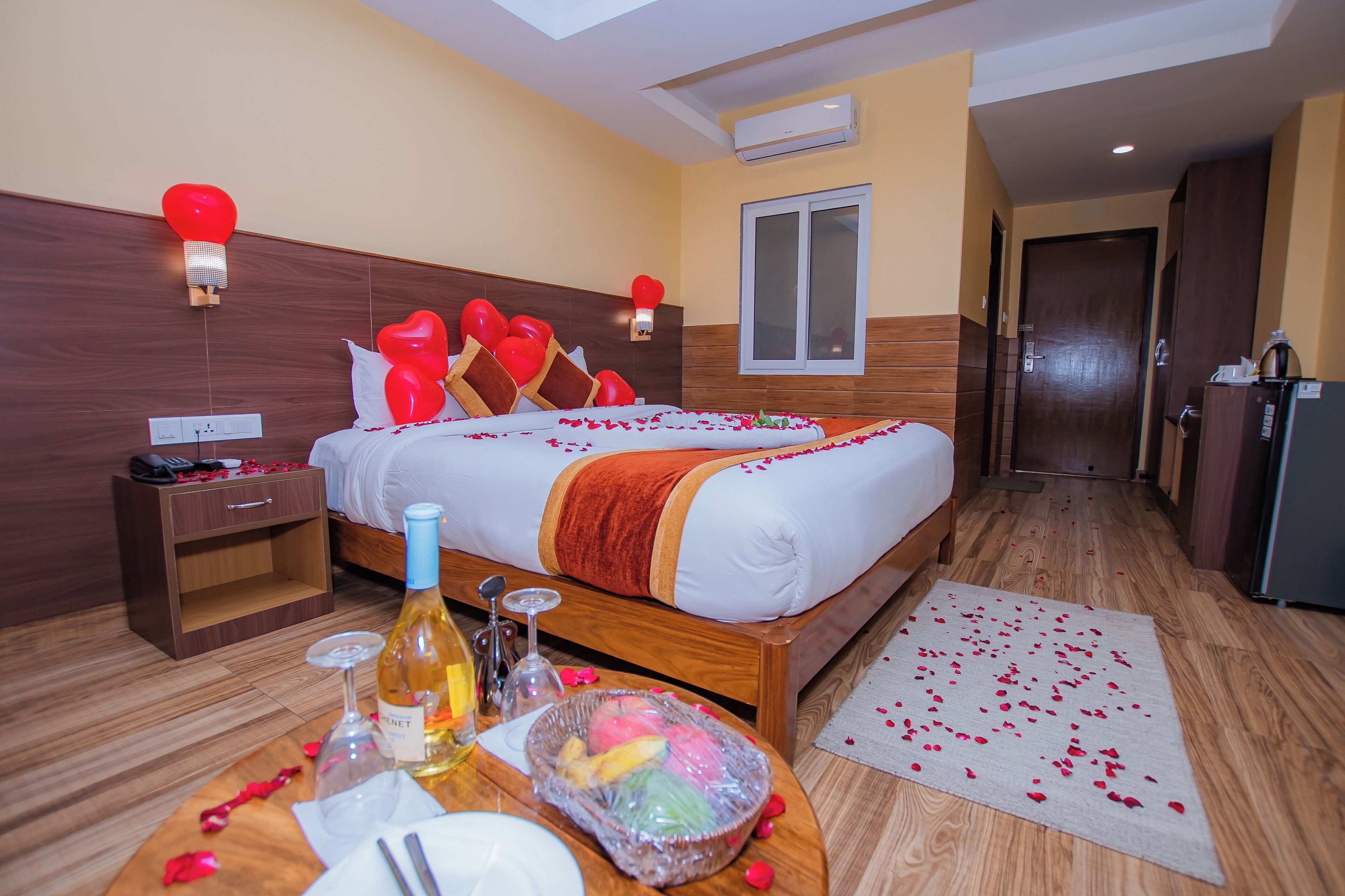 Habitación de hotel con cama, mesita de noche y una pequeña mesa con una botella, copa de vino y un frutero.
