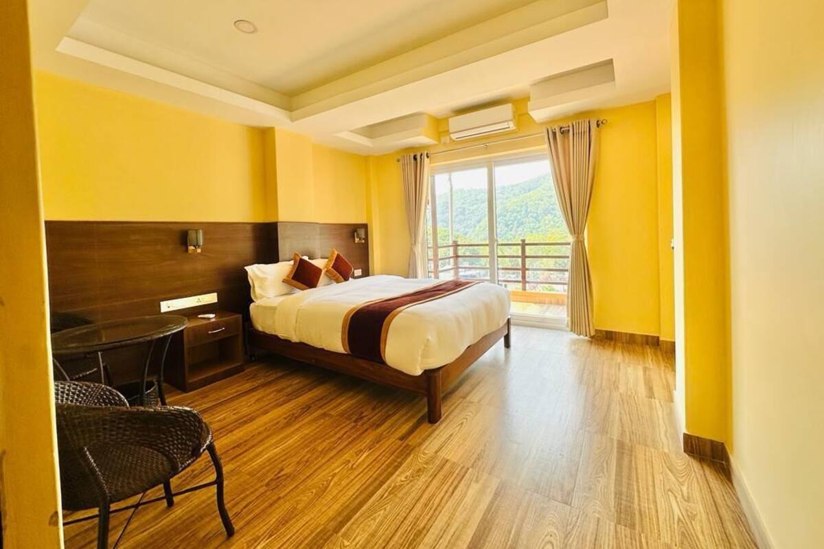 A hotel room with a wooden floor, a bed with white bedding, a desk with a chair, and a window with curtains.