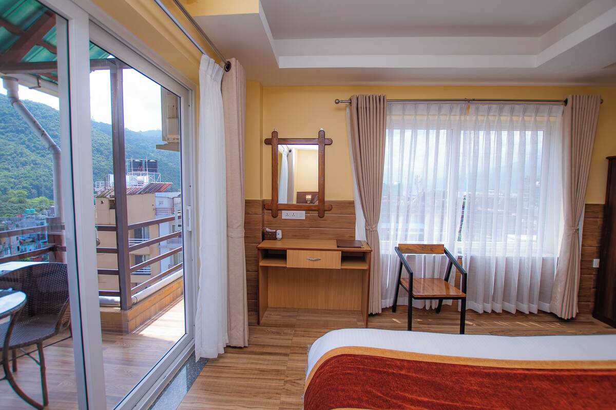 A bedroom with a wooden desk, a chair, a bed, and a balcony with a view of the mountains.