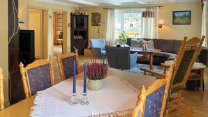 Un coin repas avec une table dressée pour quatre personnes, entourée de chaises en bois à l’assise recouverte d’un tissu à motifs floraux. Un vase contenant des fleurs roses est posé sur la table.