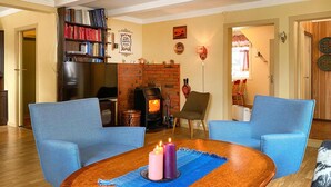 Un salon avec une cheminée en briques, deux fauteuils bleus, une table basse en bois ornée de bougies et une bibliothèque remplie de livres.