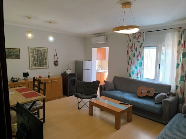 Ein Wohnzimmer mit einer Couch, einem Couchtisch, einem Esstisch mit Stühlen, einer kleinen Küchenzeile und einem Fenster mit Vorhängen.