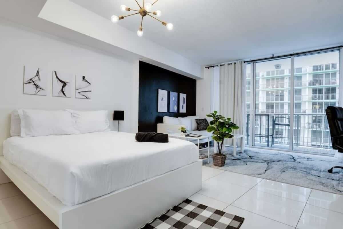A modern hotel room with a large bed, a small table, a potted plant, and a view of the city through large windows.