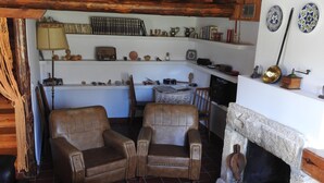 Ein gemütliches Wohnzimmer mit zwei Ledersesseln, einem Couchtisch und einem Steinkamin. Darüber befinden sich Regale mit verschiedenen Dekorationsobjekten und einer Buchsammlung.