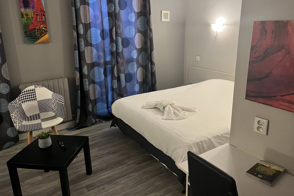 A hotel room with a bed, a small table with a plant, a chair, and a painting on the wall.